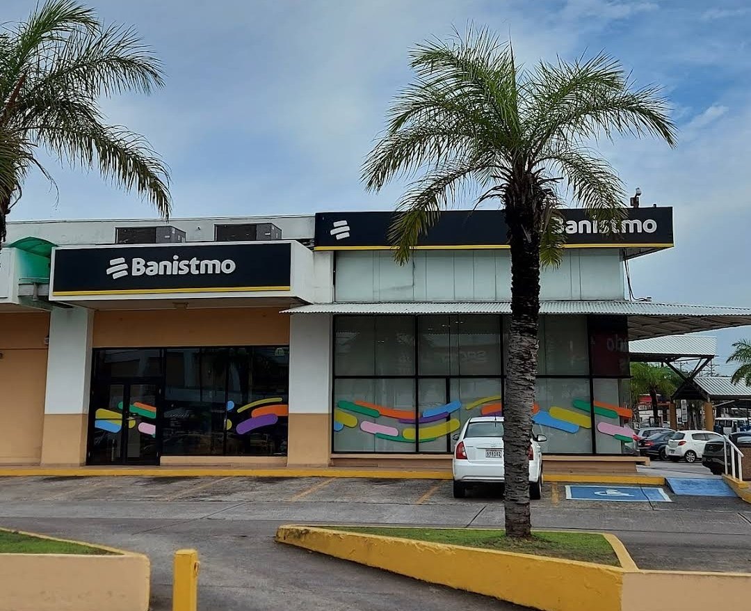 A single-storey Banistmo branch with dark signs, palm trees and a parked white car in front