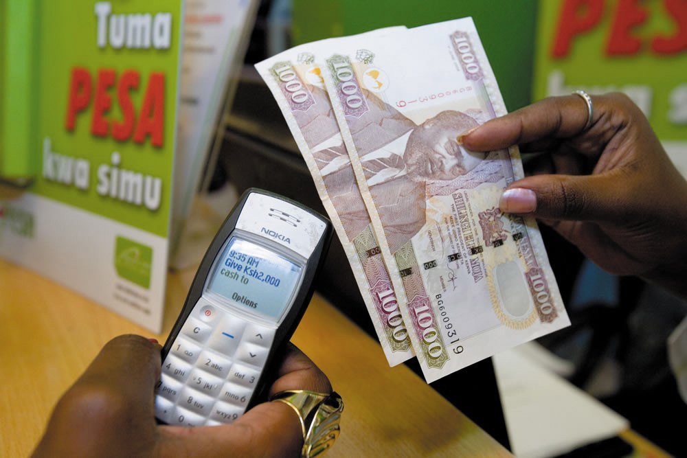 Hand holding a Nokia phone showing an M-Pesa cash message next to Kenyan 1,000-shilling banknotes
