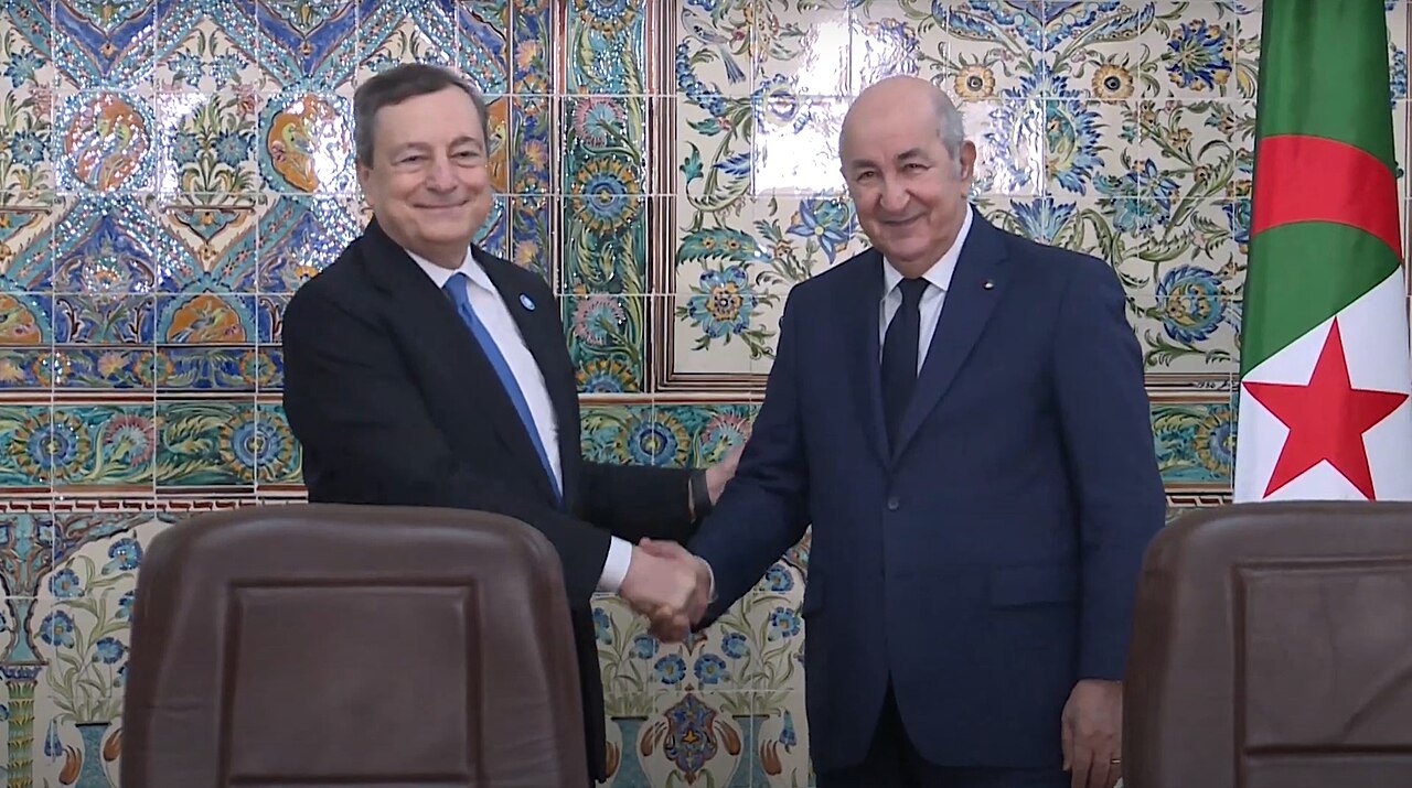 Mario Draghi and Abdelmadjid Tebboune shake hands in front of tiled wall and Algerian flag