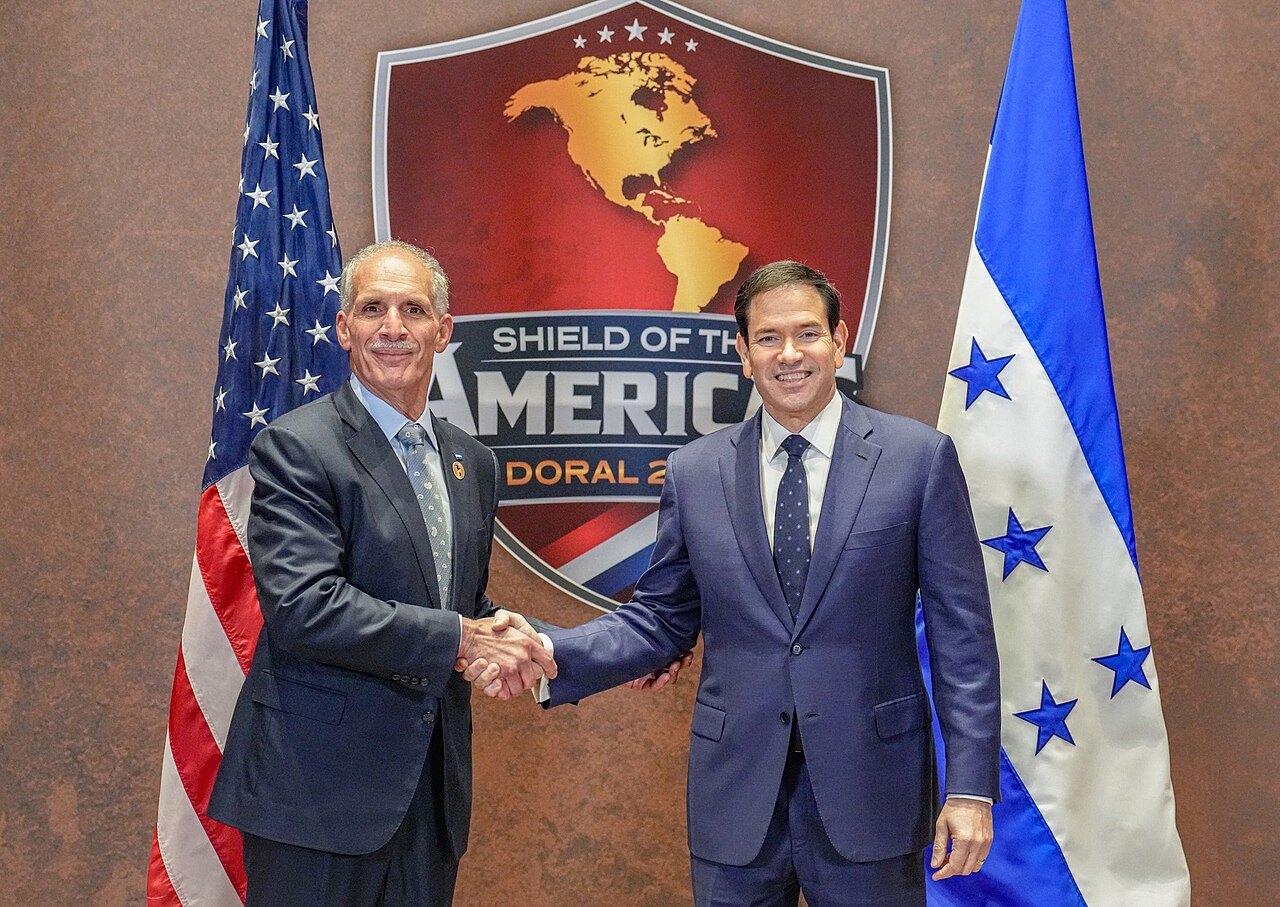 Asfura shakes hands with Marco Rubio between US and Honduran flags in front of a Shield of the Americas logo