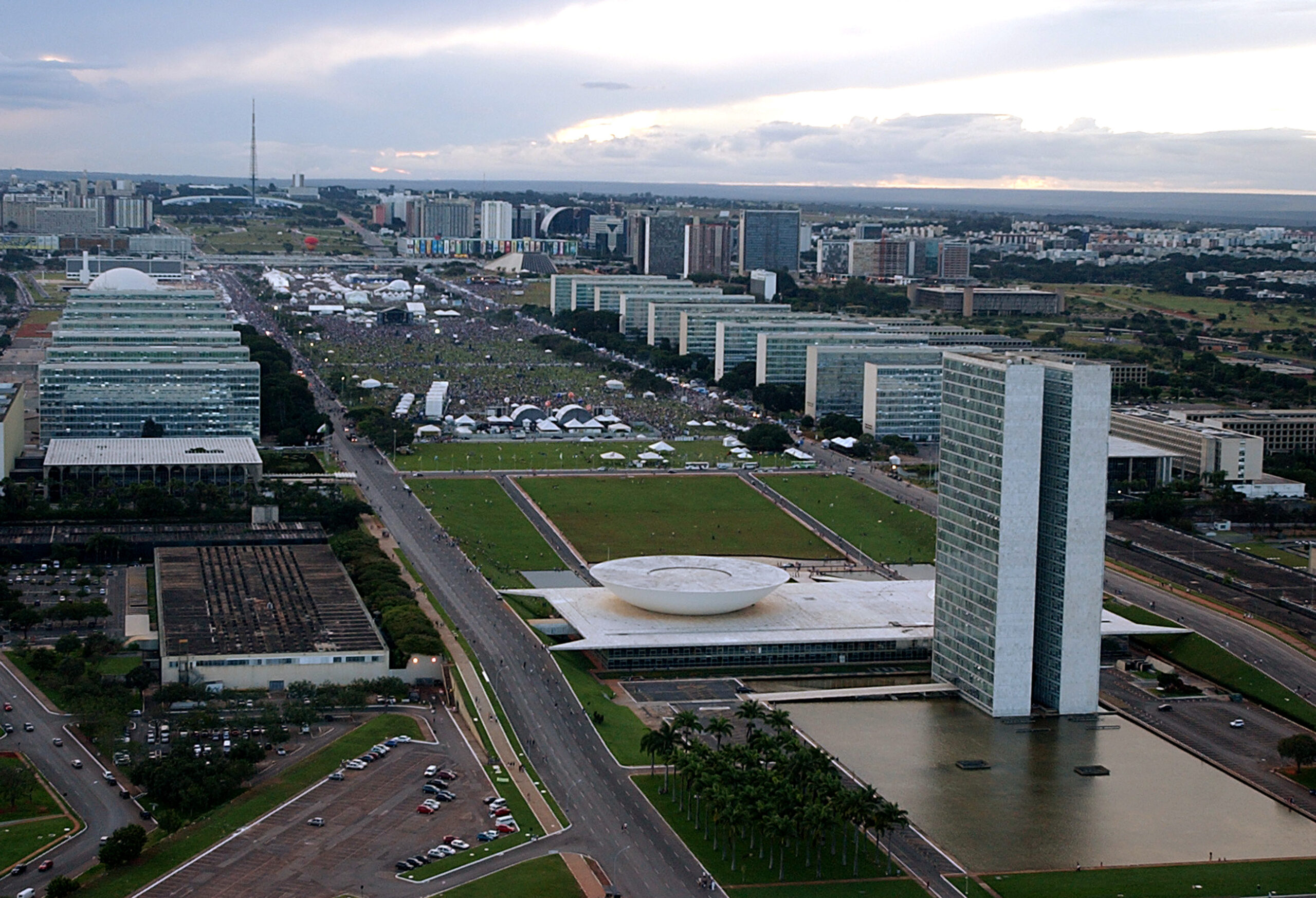 Aerial view of the National Congress tower and its bowl-shaped dome beside the ministry buildings of the Esplanada dos Ministérios in Brasília, with a large crowd gathered in the distance