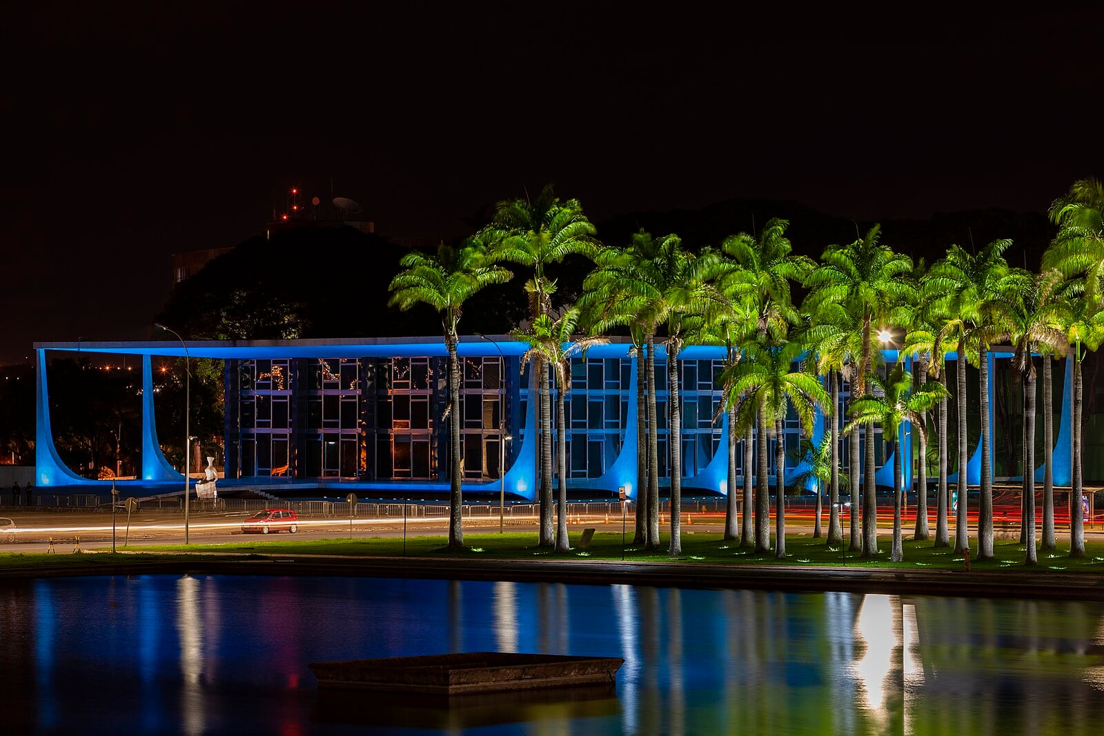 The Supreme Federal Court building in Brasília lit in blue at night, seen across a reflecting pool and palm trees