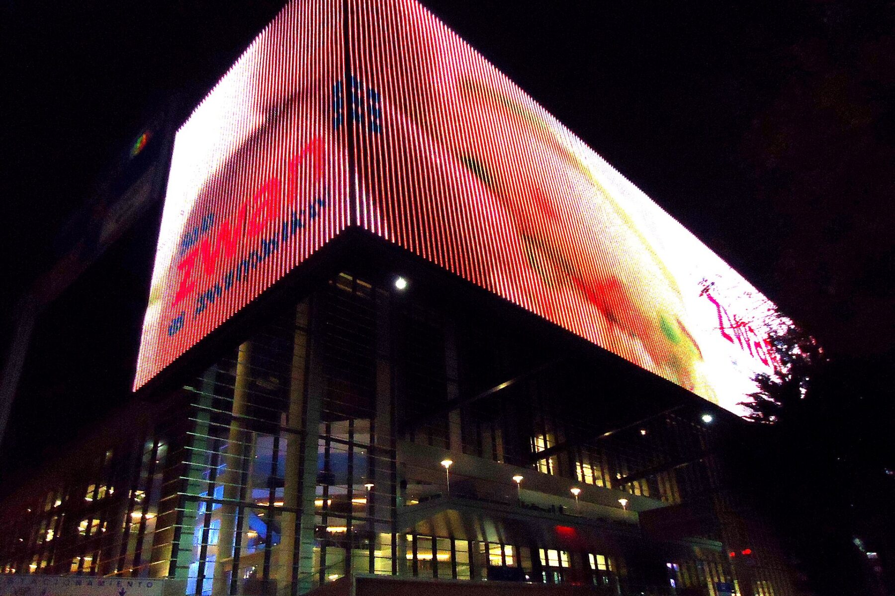 A large illuminated LED screen wrapped around the upper floors of a glass-fronted arena building at night in Mexico City