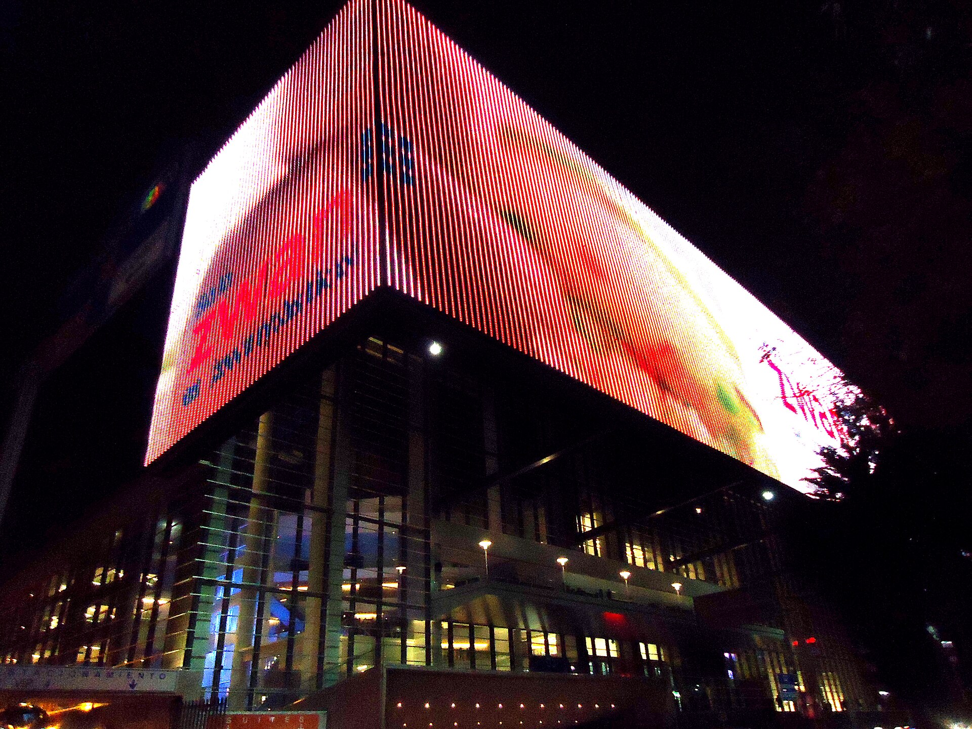 A large illuminated LED screen wrapped around the upper floors of a glass-fronted arena building at night in Mexico City