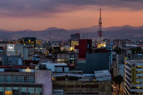Mexico City rooftops and towers at dusk with a red-orange sky and mountains on the horizon