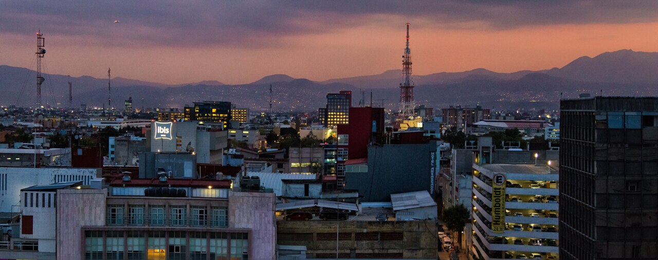 Mexico City rooftops and towers at dusk with a red-orange sky and mountains on the horizon