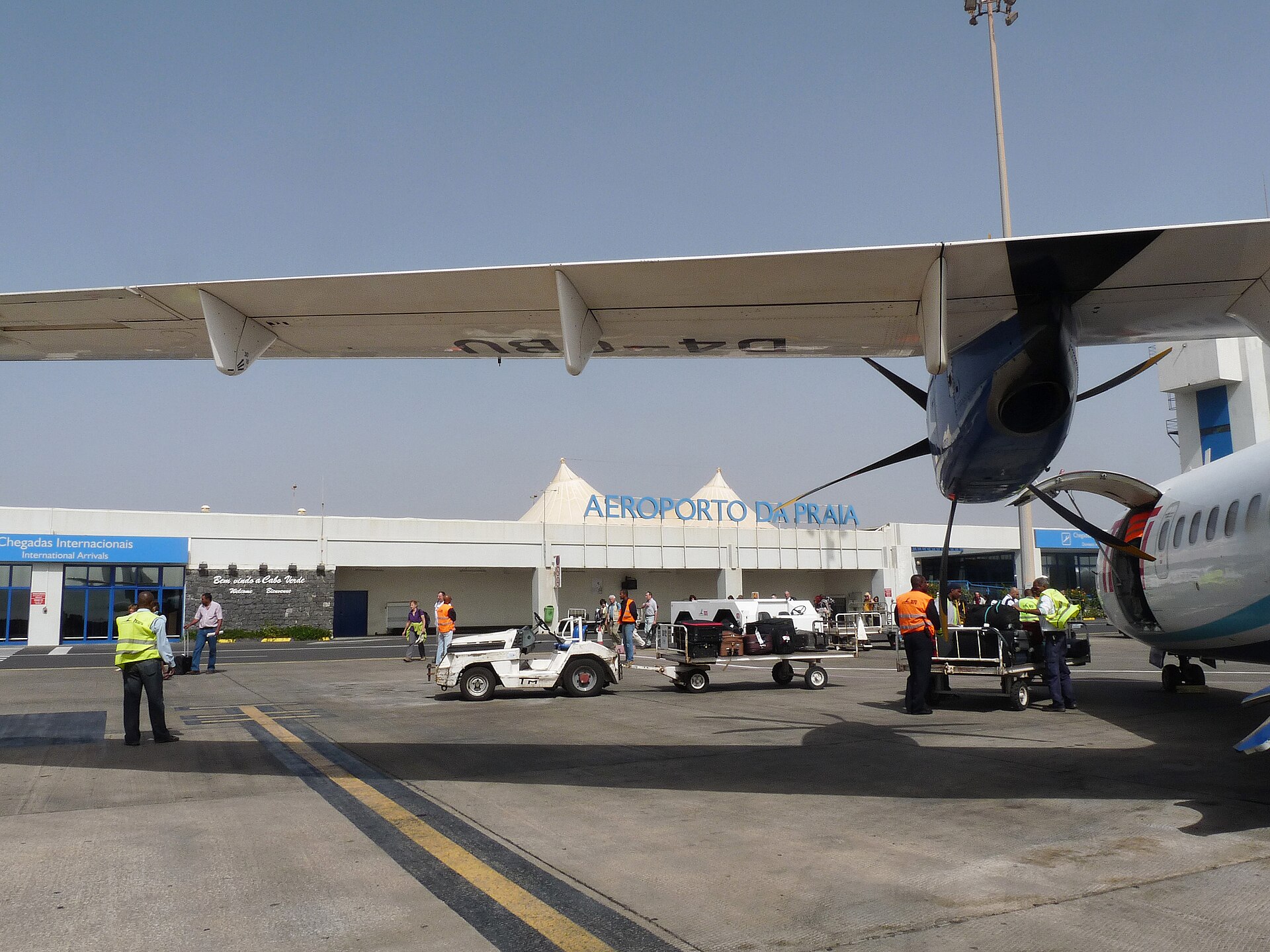 Ground staff in high-visibility vests unload luggage carts beside a turboprop wing in front of a terminal signed Aeroporto da Praia