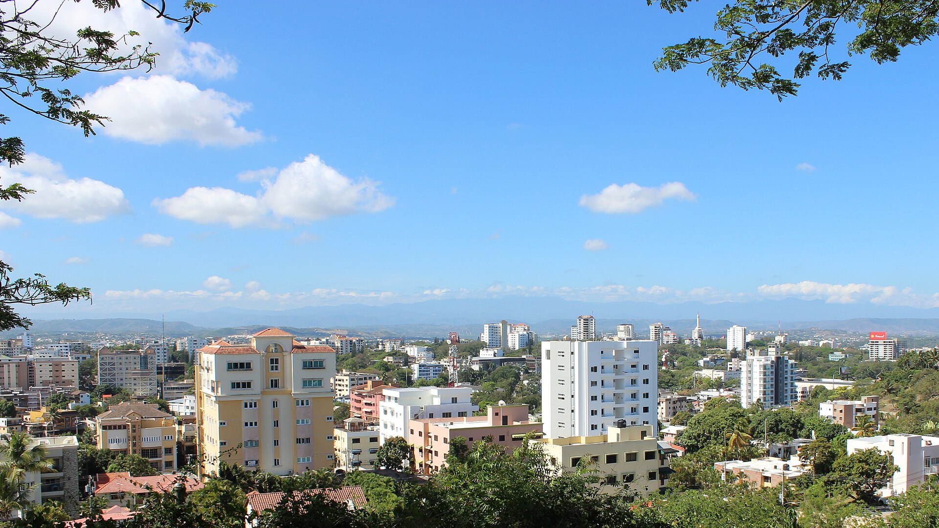 View over the rooftops and trees of Santiago de los Caballeros under a blue sky, with hills on the horizon