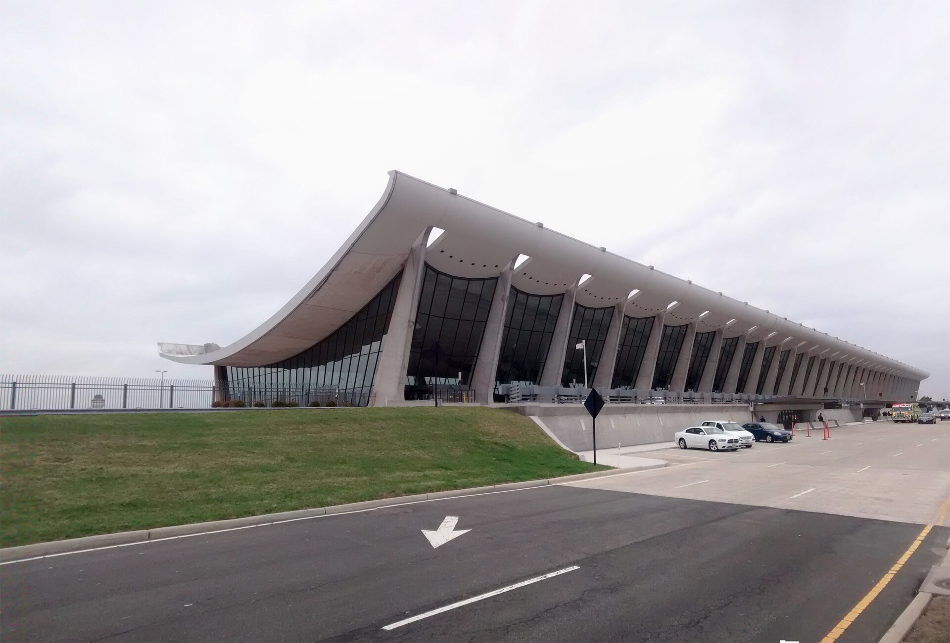 The curved concrete roof and tall glass facade of the main terminal at Washington Dulles International Airport under a grey sky, with parked cars in the foreground
