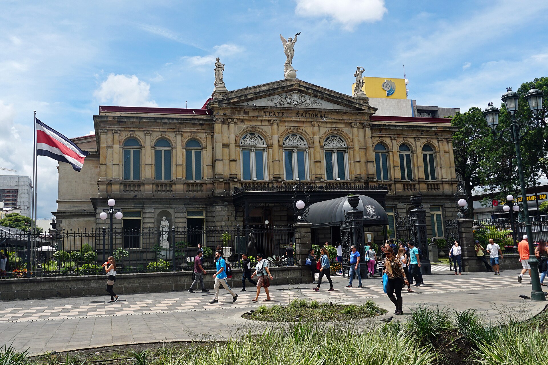 The neoclassical facade of the National Theatre in San Jose, Costa Rica, with pedestrians in the plaza in front, a Costa Rican flag and statues on the roofline