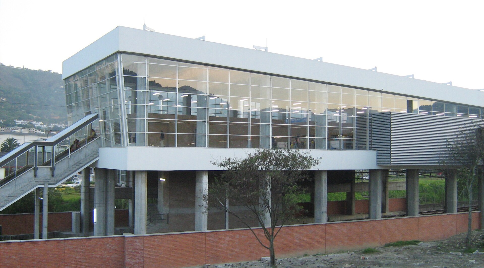 A glass-fronted metro station building on concrete pillars with a staircase on the left and green hills behind