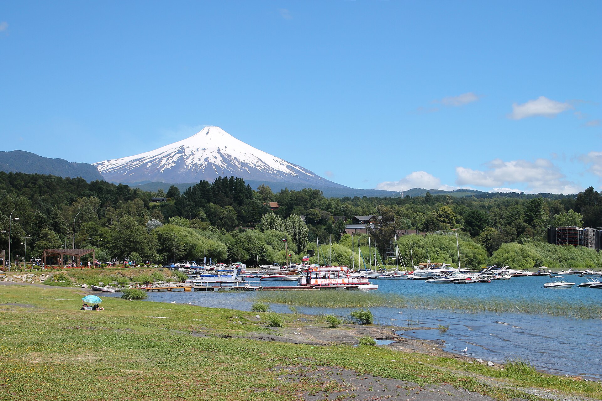 Snow-capped Villarrica volcano above forest and the shore of Lake Villarrica at Pucón on a sunny day, with boats moored near the beach