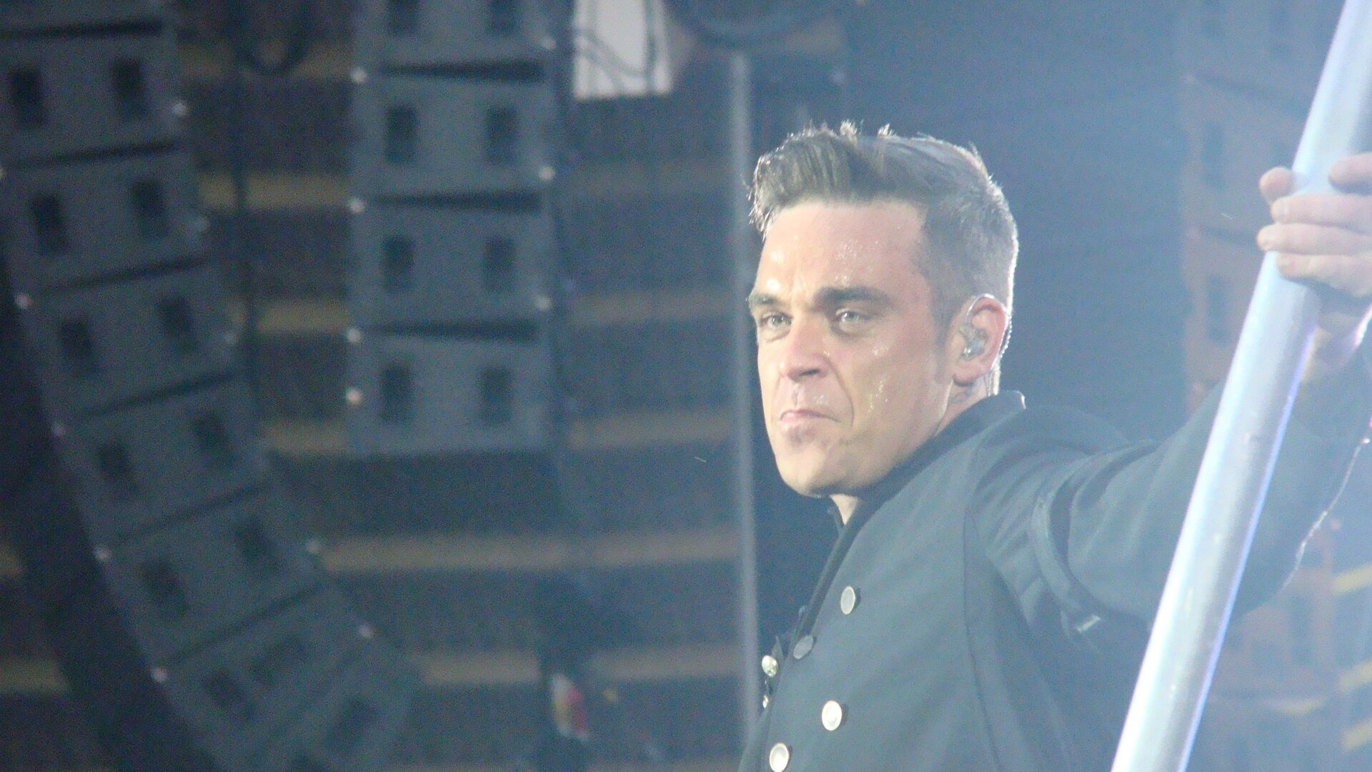 Robbie Williams in a dark military-style jacket holds a metal pole on stage in front of stacked loudspeakers