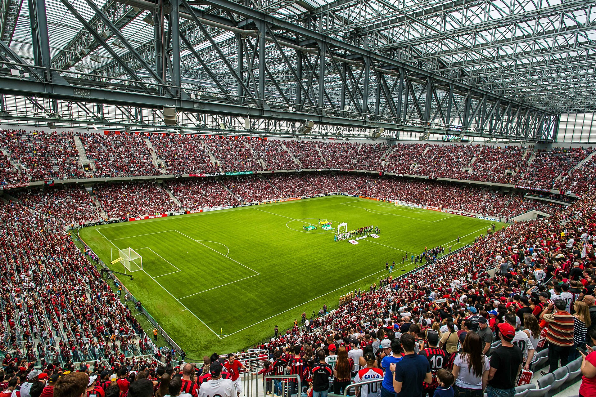 Interior of Arena da Baixada in Curitiba during a football match, with packed stands under a steel and glass roof around a green pitch
