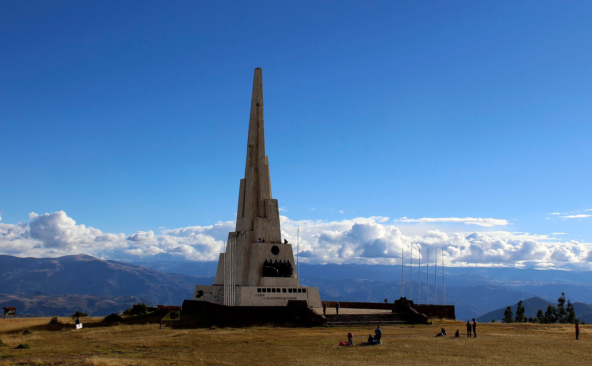 A tall white stone obelisk on a grassy plateau under a deep blue sky, with Andean ridges and clouds behind and a few visitors sitting on the grass
