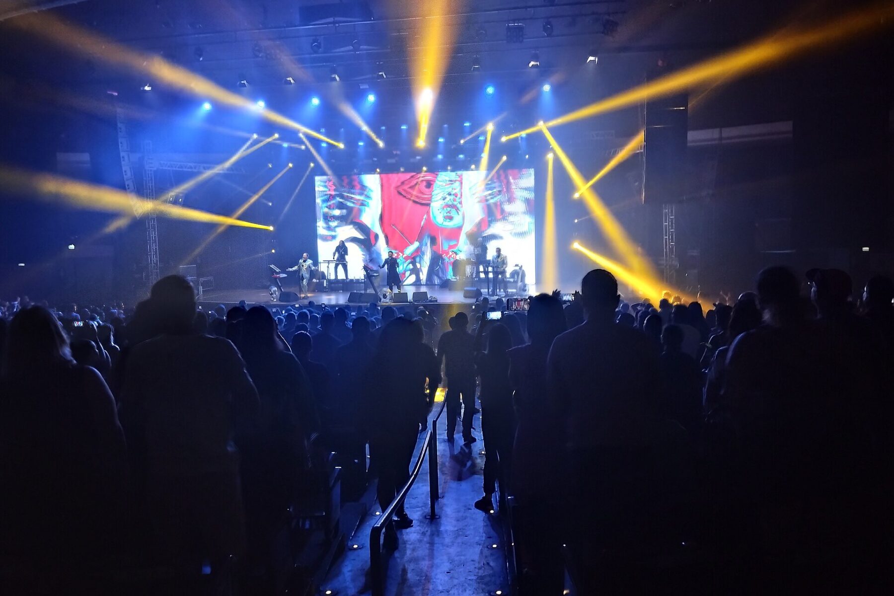 Crowd facing a lit stage with a video screen and yellow beams inside the Auditório Araújo Vianna in Porto Alegre during a December 2025 concert