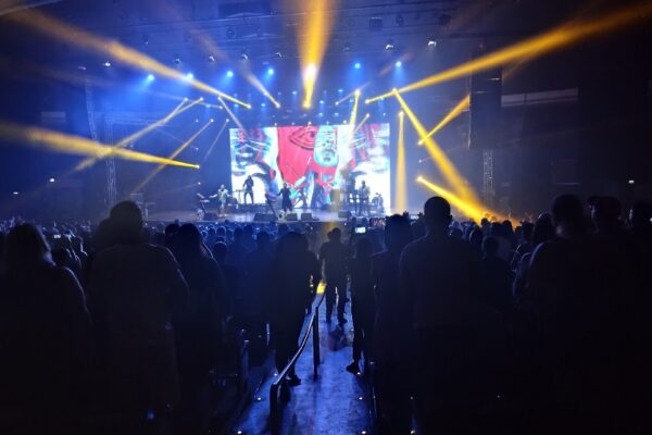 Crowd facing a lit stage with a video screen and yellow beams inside the Auditório Araújo Vianna in Porto Alegre during a December 2025 concert