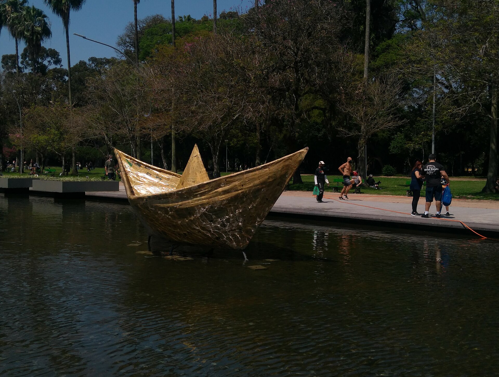 A golden paper-boat sculpture on a pond in the Parque da Redenção in Porto Alegre, with trees and walkers behind