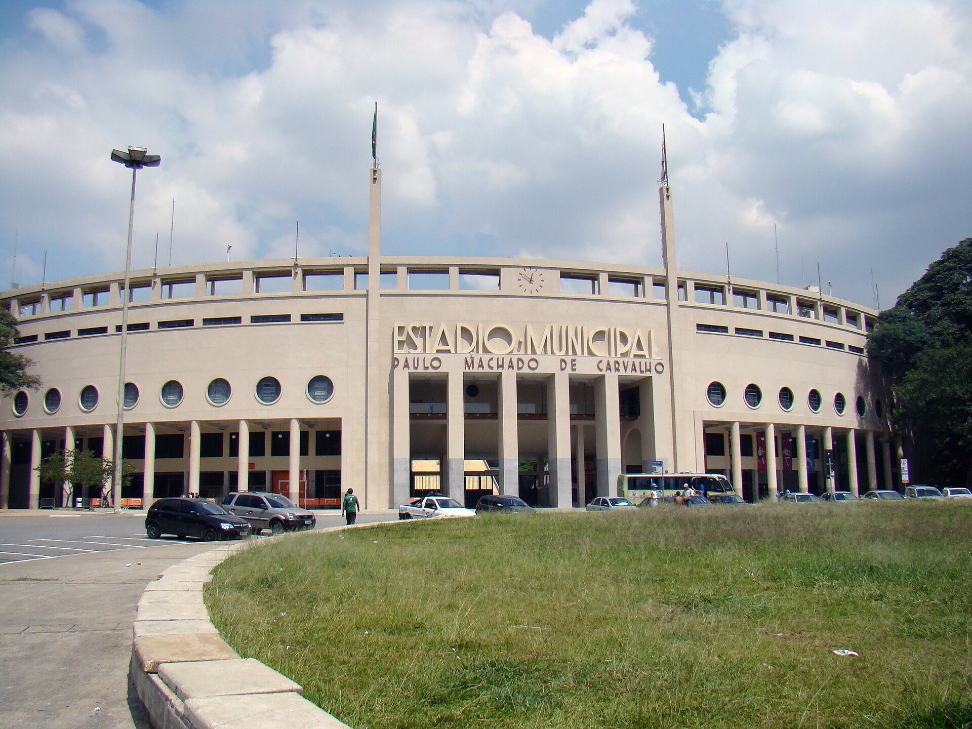 The curved cream-coloured facade of the Pacaembu stadium in São Paulo, with the words Estádio Municipal Paulo Machado de Carvalho above the entrance and a few cars parked in front