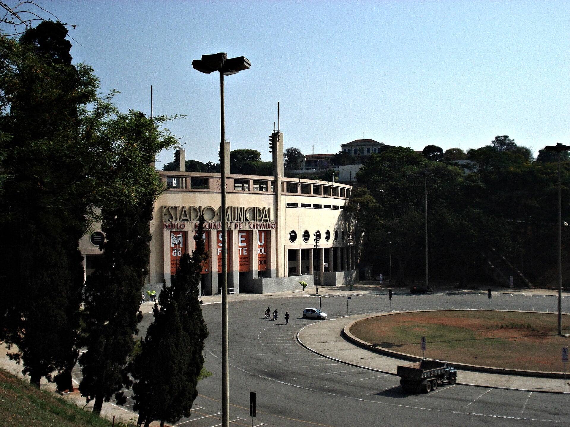 The Pacaembu stadium facade seen from a slope above Praça Charles Miller, with tall trees in the foreground and a road circling a round island in front