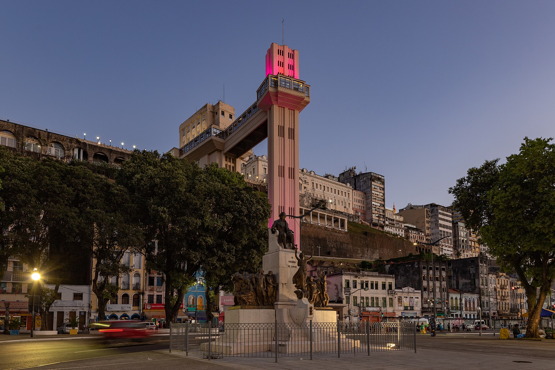 The Elevador Lacerda tower in Salvador at dusk, lit pink, above a monument and a street