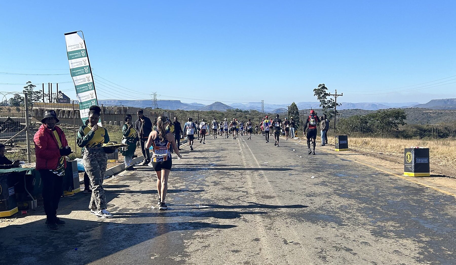 Runners on a road in KwaZulu-Natal during the 2024 Comrades Marathon, with spectators and a refreshment point on the left and hills behind