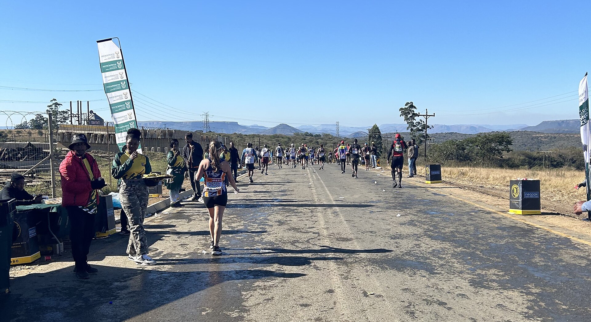Runners on a road in KwaZulu-Natal during the 2024 Comrades Marathon, with spectators and a refreshment point on the left and hills behind