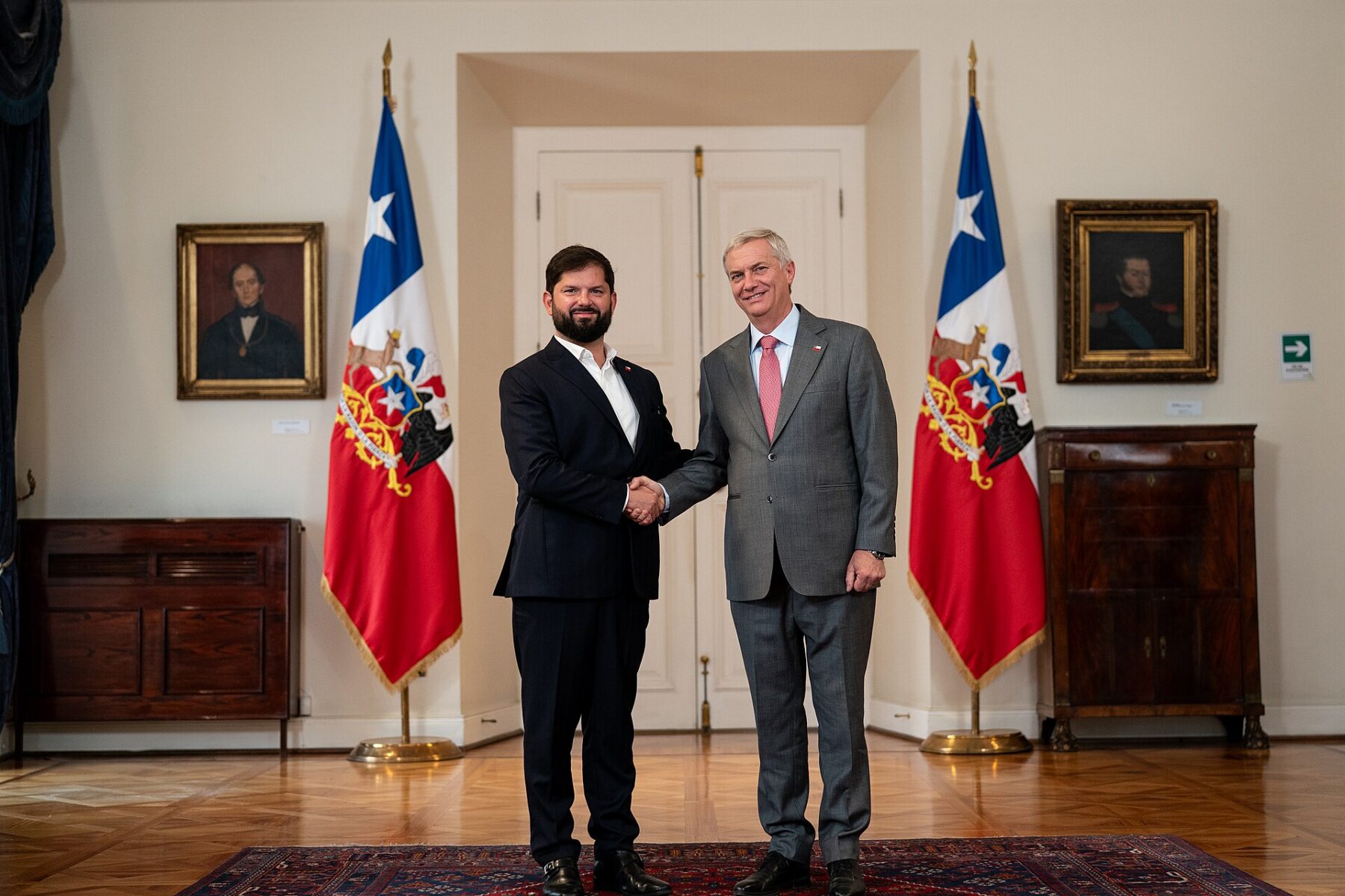 Two men in suits shake hands between two Chilean flags in a formal room with framed portraits, a white double door and a patterned rug