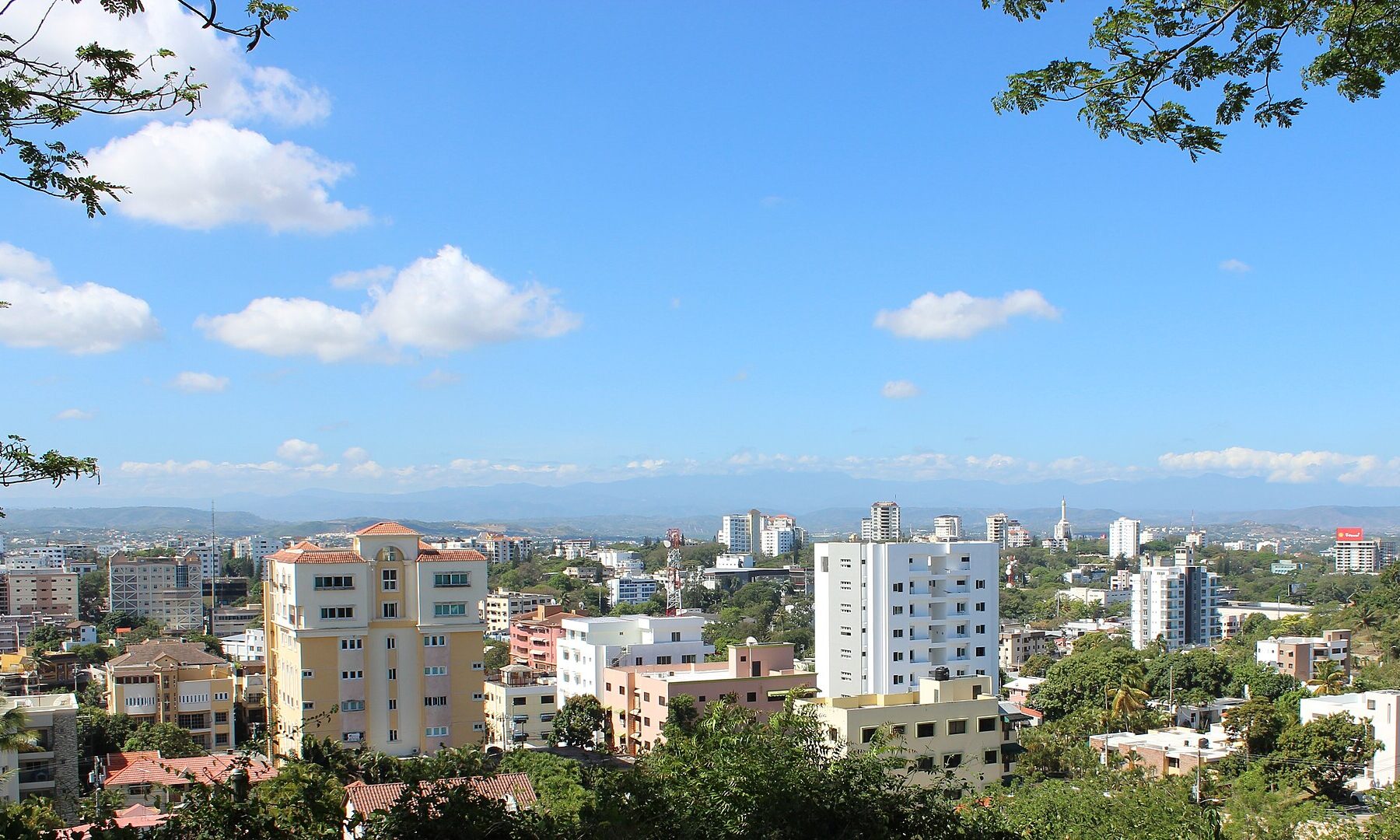Hillside view over Santiago de los Caballeros, with apartment towers among trees, mountains on the horizon and the white Monument to the Heroes of the Restoration right of centre