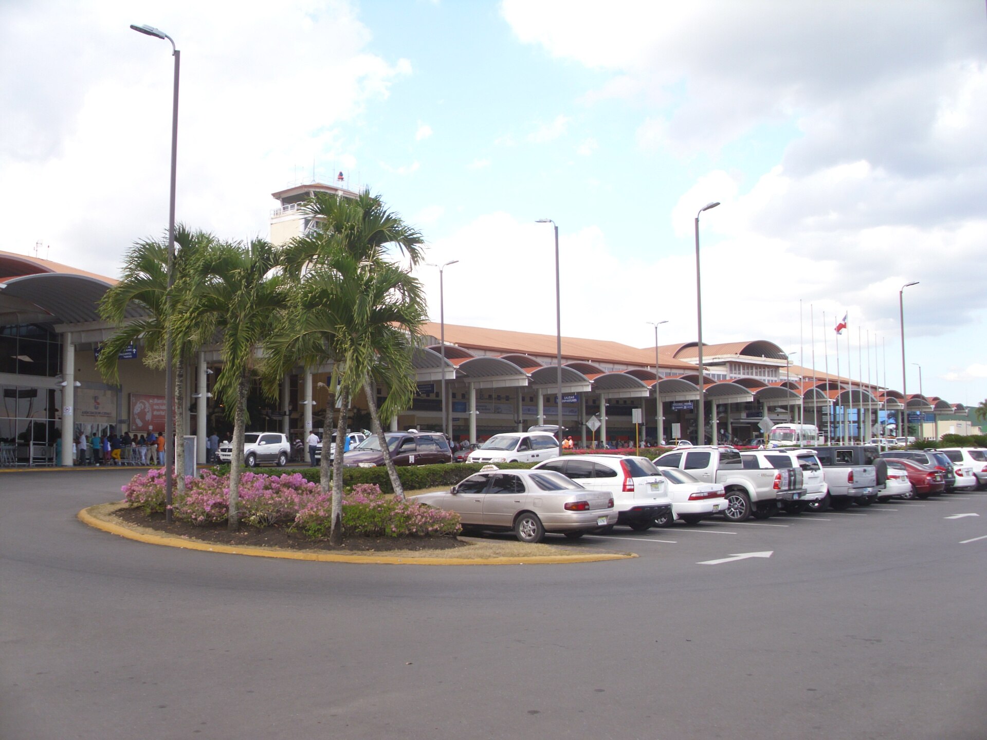 Low terminal building with orange tiled roofs and a control tower, palm trees and a flowerbed in front, and cars parked in the open-air car park