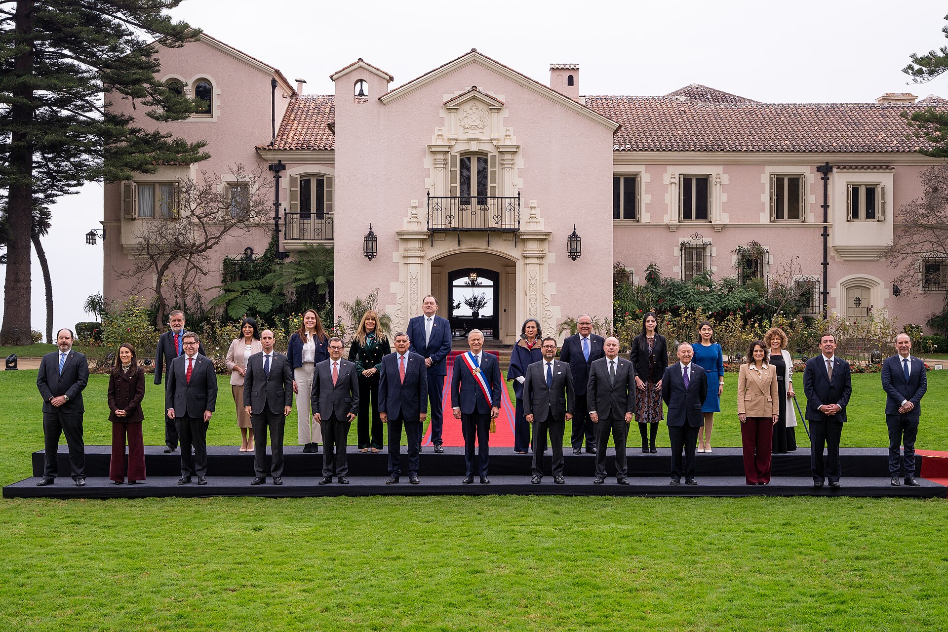 A group of about two dozen men and women in formal clothes stands on a low stage on a lawn in front of a pink mansion, with a man wearing a presidential sash in the centre