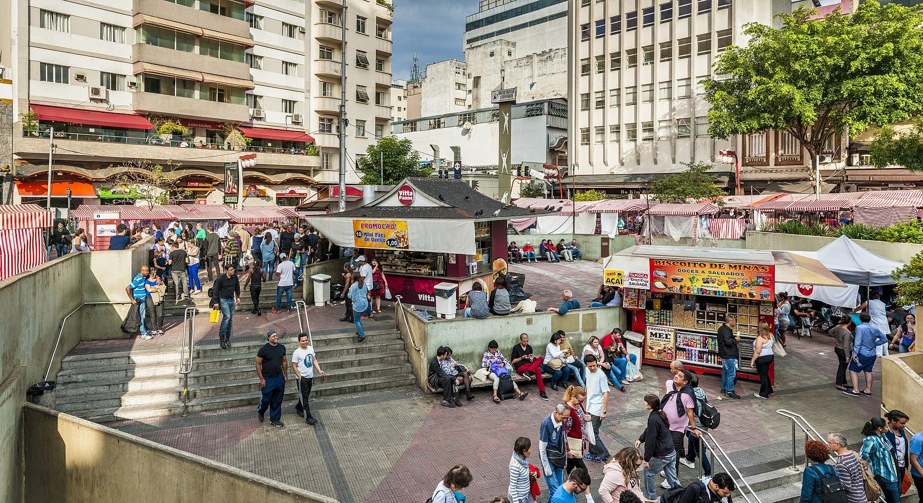 Pedestrians cross a brick plaza with stairs down to a metro entrance and food and juice stalls, with apartment blocks behind, in São Paulo's Liberdade district