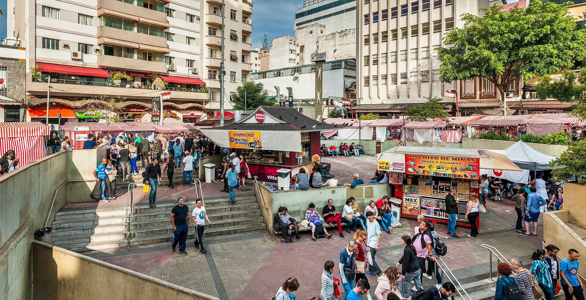 Pedestrians cross a brick plaza with stairs down to a metro entrance and food and juice stalls, with apartment blocks behind, in São Paulo's Liberdade district