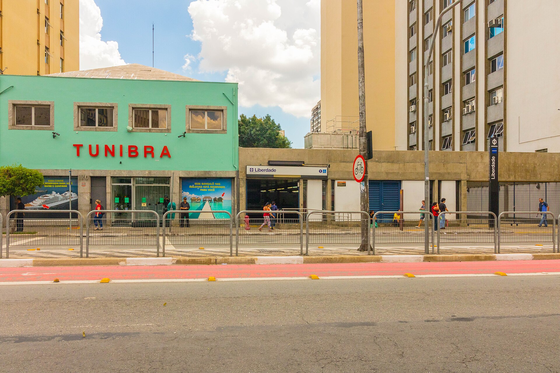 A green low building with a red travel agency sign beside the Liberdade metro station entrance on a São Paulo avenue, with a bike lane in the foreground