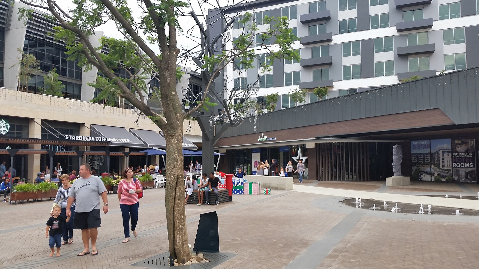 Shoppers walk through a paved square at Menlyn Maine shopping centre in Pretoria, with cafes and apartment buildings around it