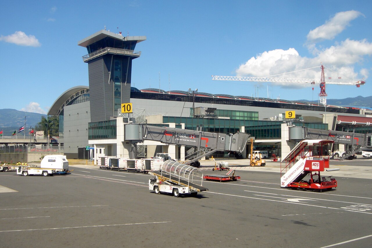 Airport terminal with a tall control tower, jet bridges at gates 9 and 10 and ground service vehicles on the apron under a blue sky