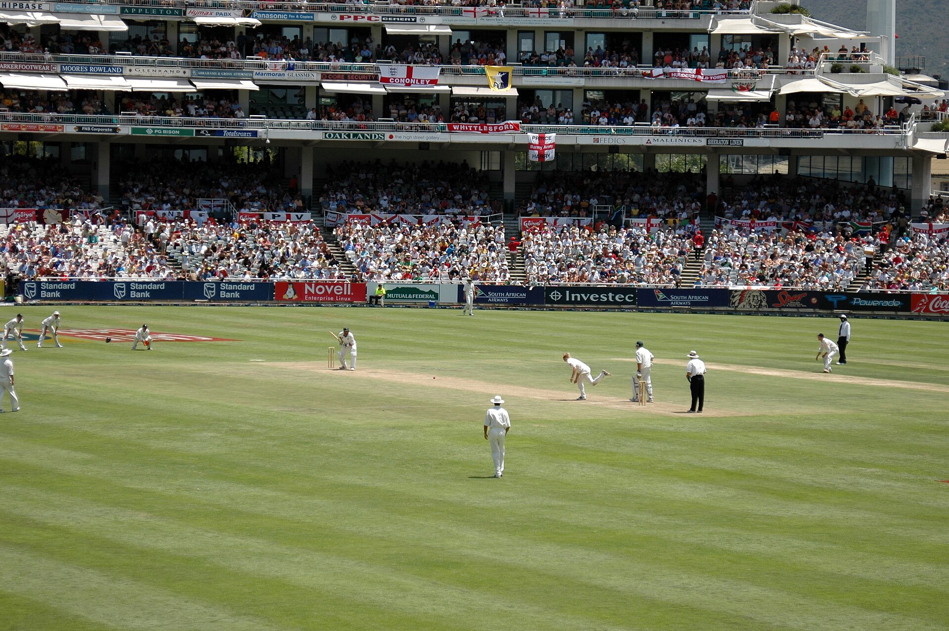A Test match in progress at Newlands in Cape Town, with a bowler delivering to a batter, fielders in white on a green outfield and full stands hung with England flags
