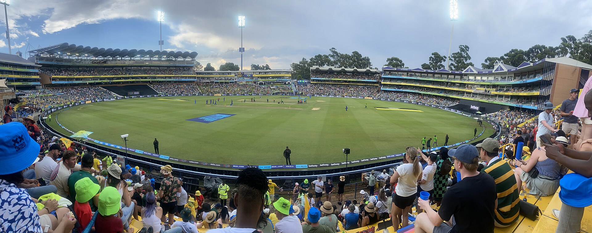 Panoramic view of the Wanderers Stadium in Johannesburg from the stands, with a crowd in the foreground, floodlight towers and cricketers on a green field