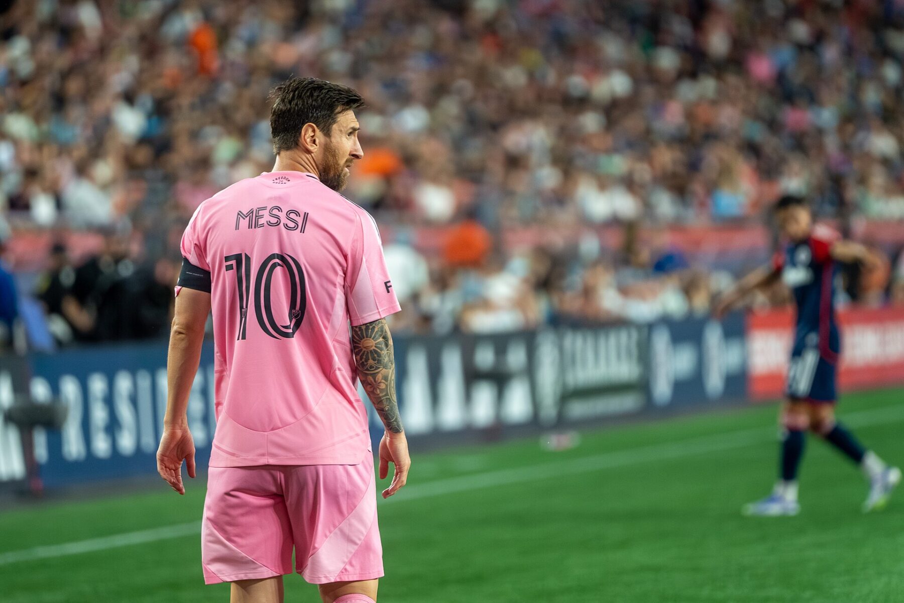 A footballer in a pink Inter Miami shirt with the name Messi and the number 10 stands on the pitch, seen from behind, with a crowd in the stands