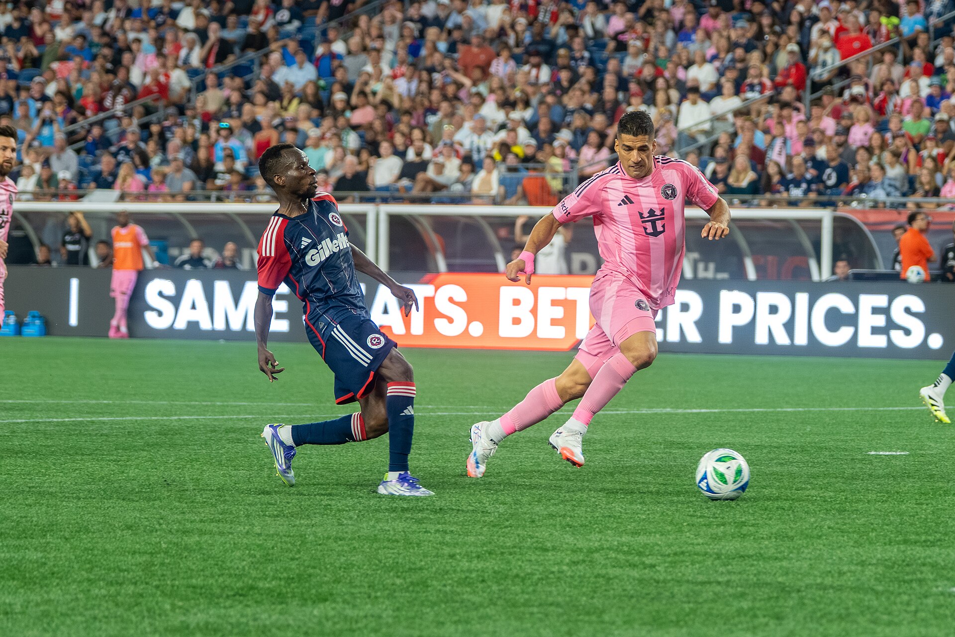 A footballer in a pink Inter Miami kit runs with the ball past a New England Revolution player in a navy kit, in front of a full stand
