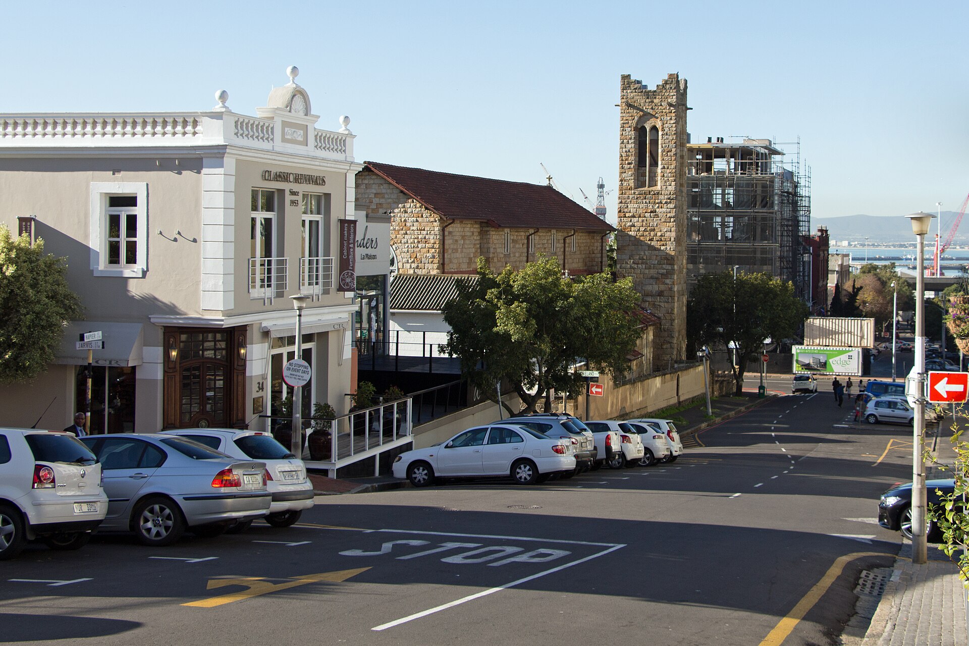 A hillside street in De Waterkant, Cape Town, with a cream corner building, a stone church tower under repair and parked cars