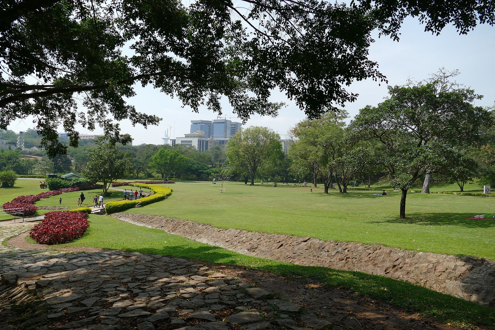 A lawn with flower beds and trees in Millennium Park in Abuja, with city buildings in the distance