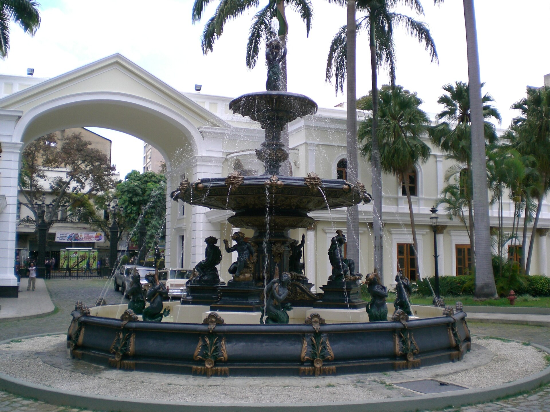 A dark ornate tiered fountain with sculpted figures in a courtyard with palm trees, a white arched gateway and cream colonial buildings