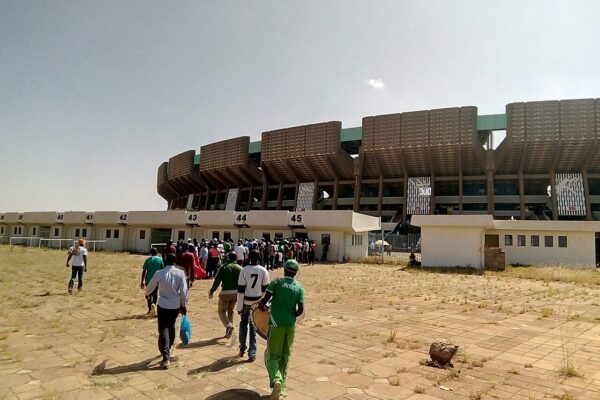 Fans walk across a paved forecourt toward the curved concrete wall and entrance gates of Kasarani Stadium in Nairobi under a hazy sky