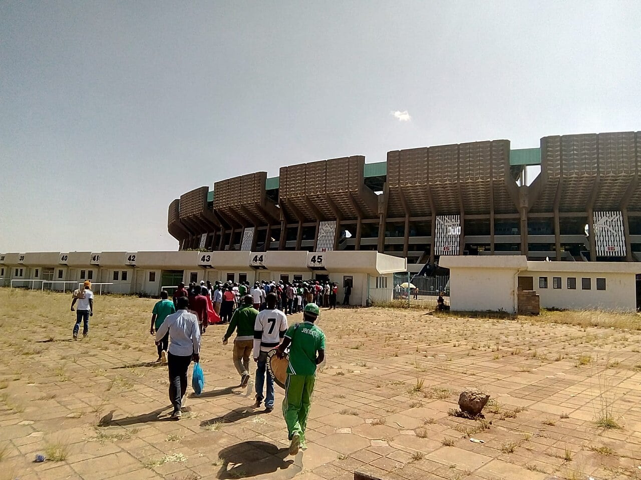 Fans walk across a paved forecourt toward the curved concrete wall and entrance gates of Kasarani Stadium in Nairobi under a hazy sky