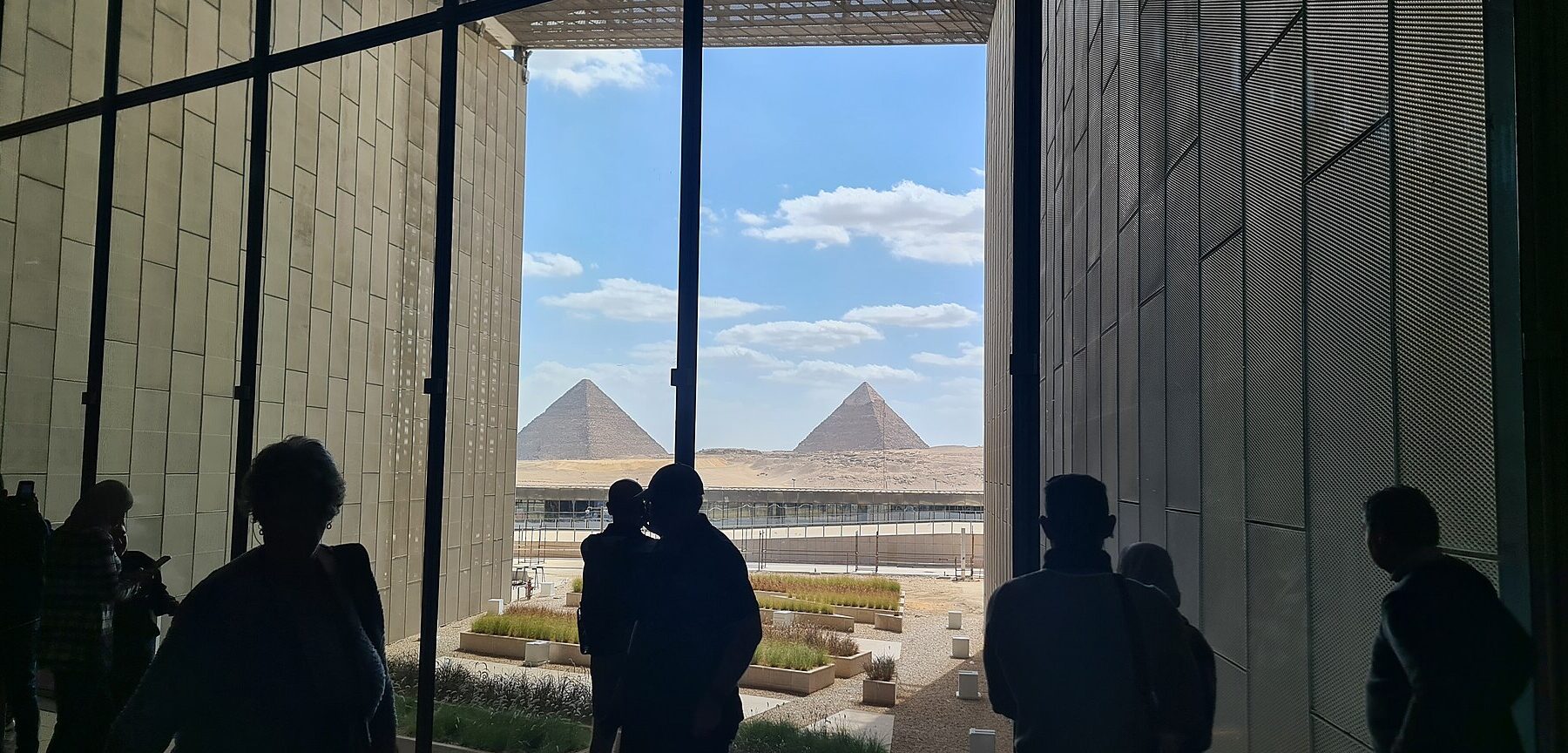 Visitors in silhouette look out from the Grand Egyptian Museum between two stone walls at the Pyramids of Giza under a blue sky