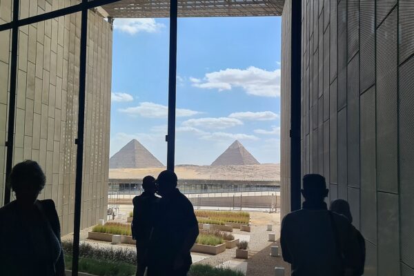Visitors in silhouette look out from the Grand Egyptian Museum between two stone walls at the Pyramids of Giza under a blue sky