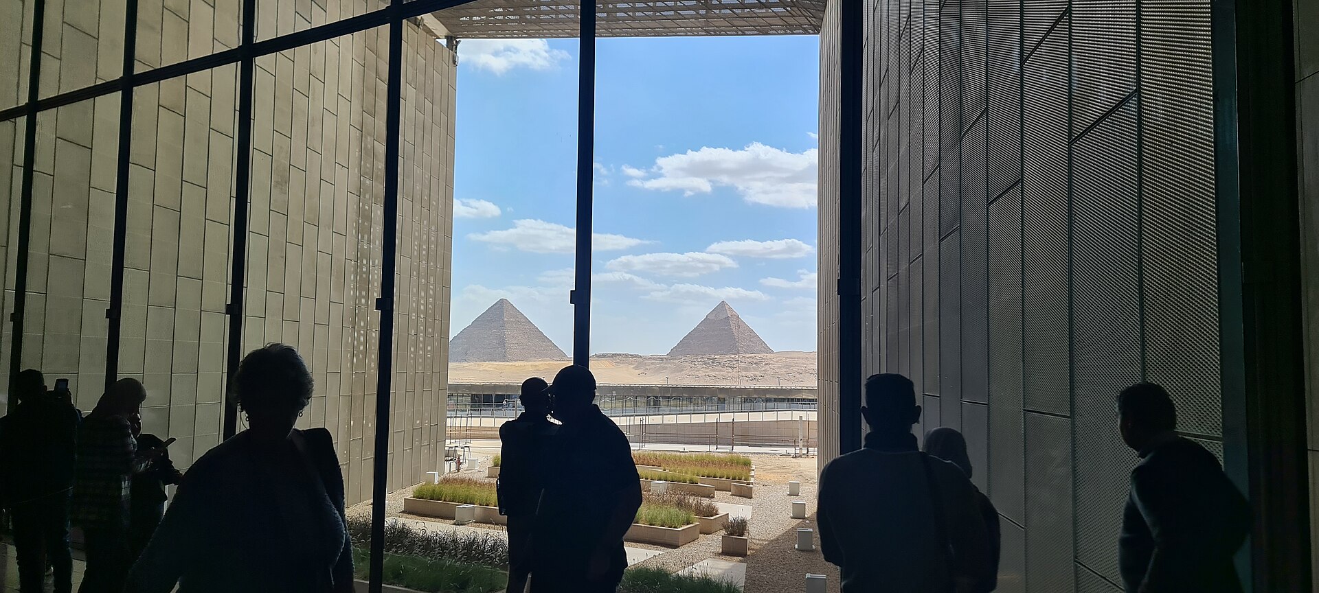 Visitors in silhouette look out from the Grand Egyptian Museum between two stone walls at the Pyramids of Giza under a blue sky