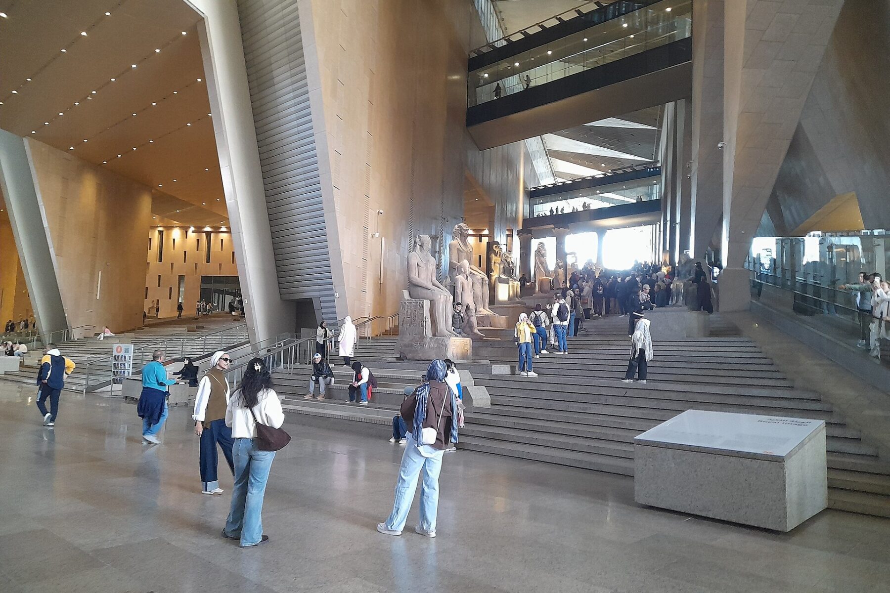 Visitors stand and sit on the wide Grand Stairs in the high Grand Hall of the Grand Egyptian Museum beside rows of ancient statues
