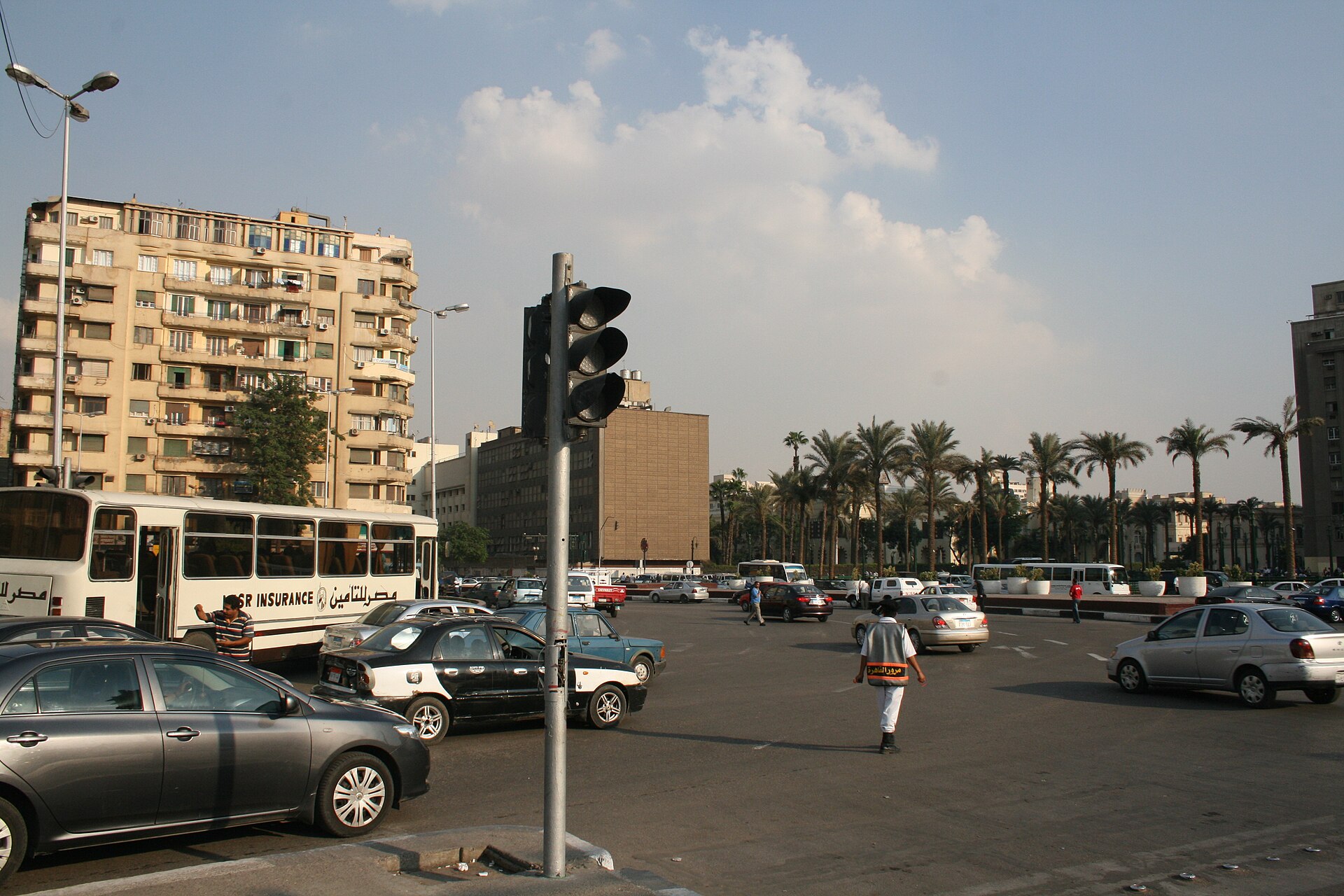 Cars, a white bus and pedestrians on a wide square lined with palm trees and apartment blocks under a partly cloudy sky, with a traffic light in the foreground